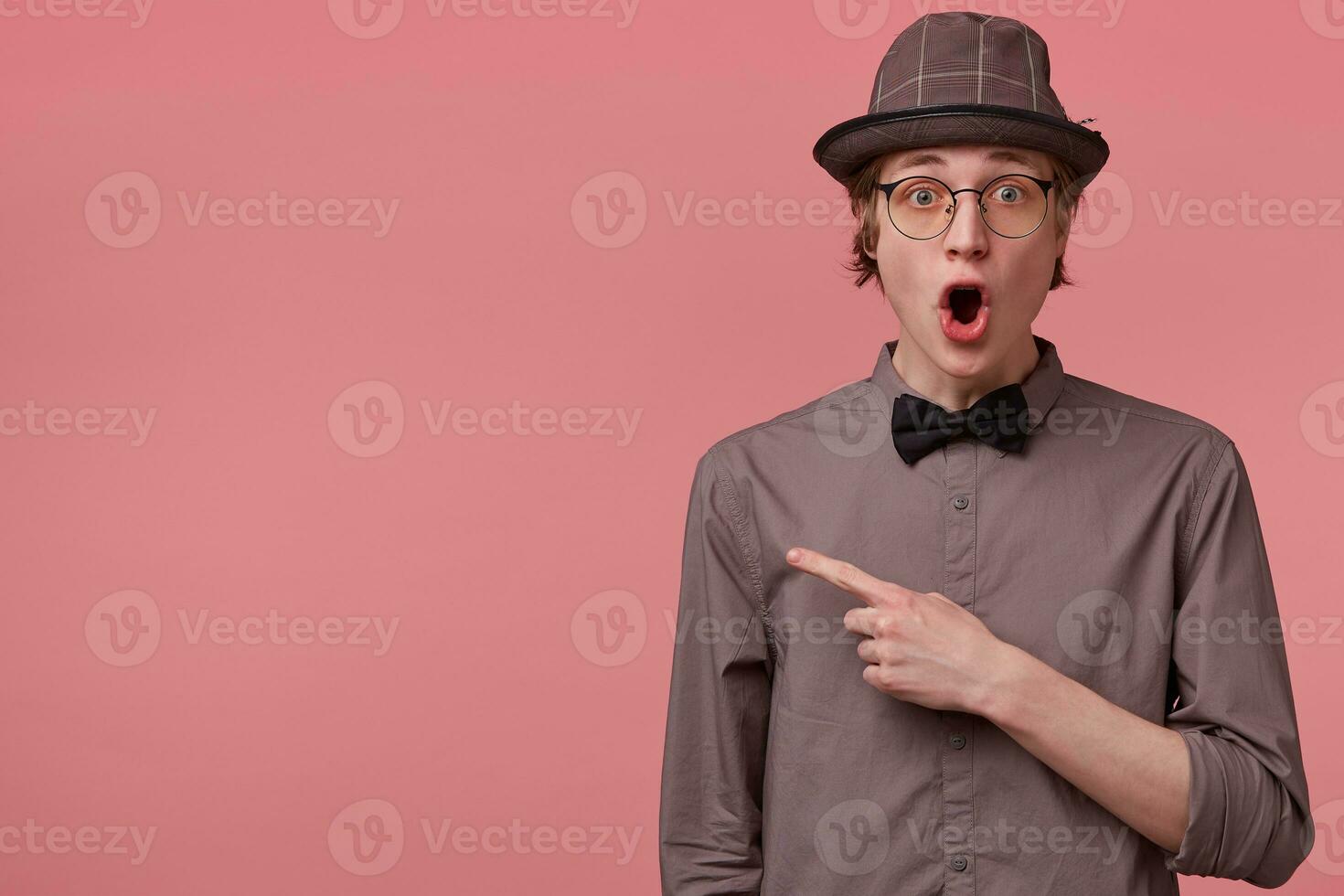 Surprised elegant young slim guy opened his mouth in amazement, is overwhelmed with emotions shocked isolated on pink, pointing index finger to the left on copy space, omg photo