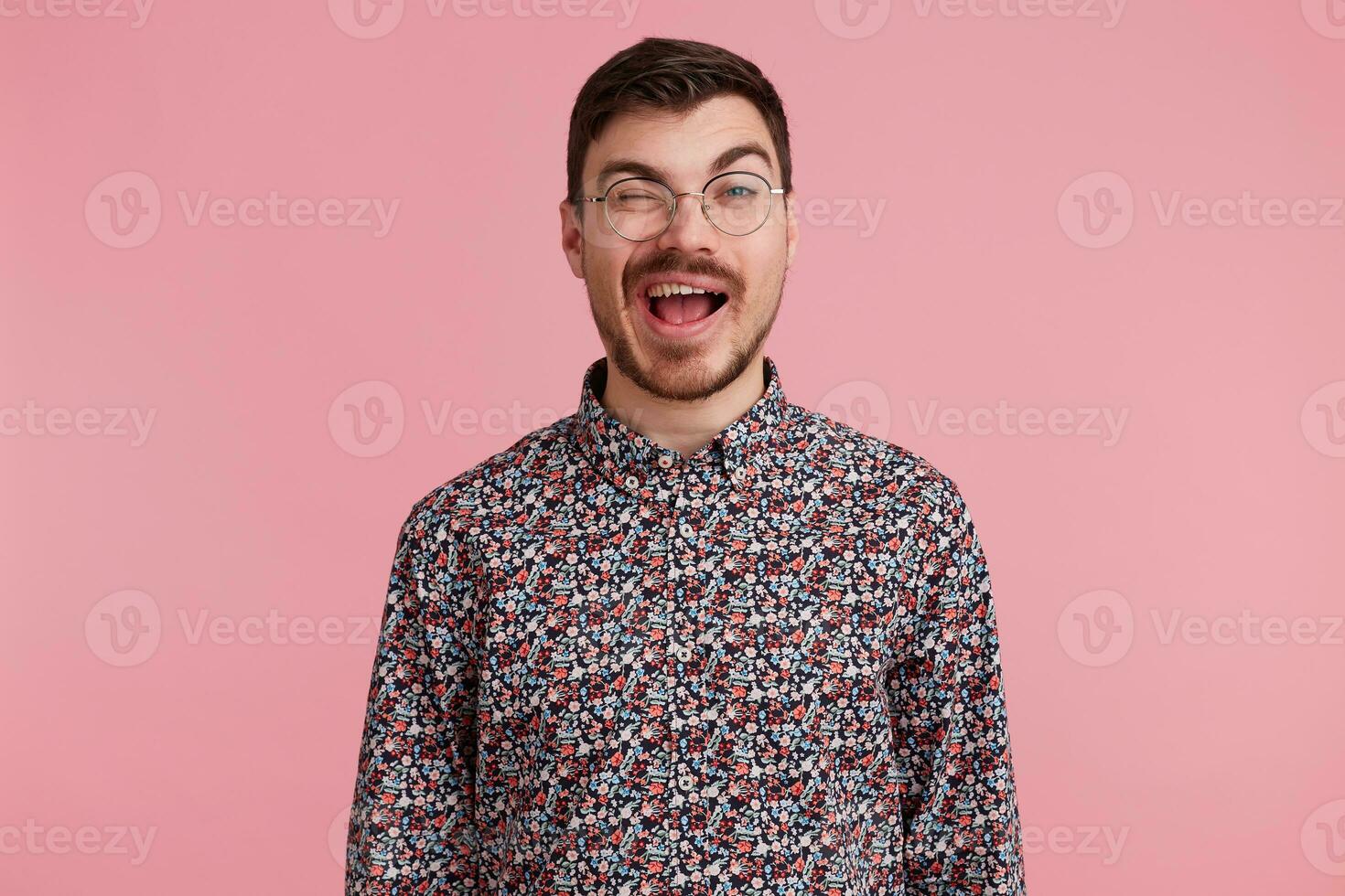 Close up photo of sweet heartbreaking darkhaired attractive man in glasses, flirting with someone, winks showing approval predisposed interes, in coloful shirt, isolated over pinkstudio background