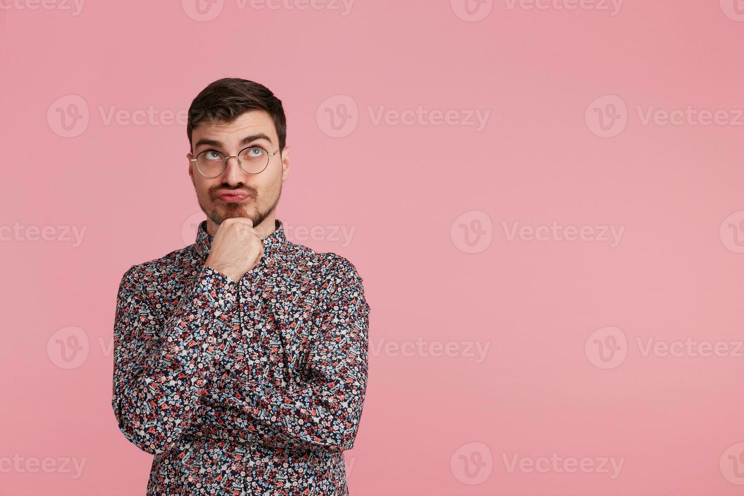 Young man in colorful shirt looking upwards, copy space on the right side,having doubts and with confuse face expression while scratching his chin isolated on pink background photo