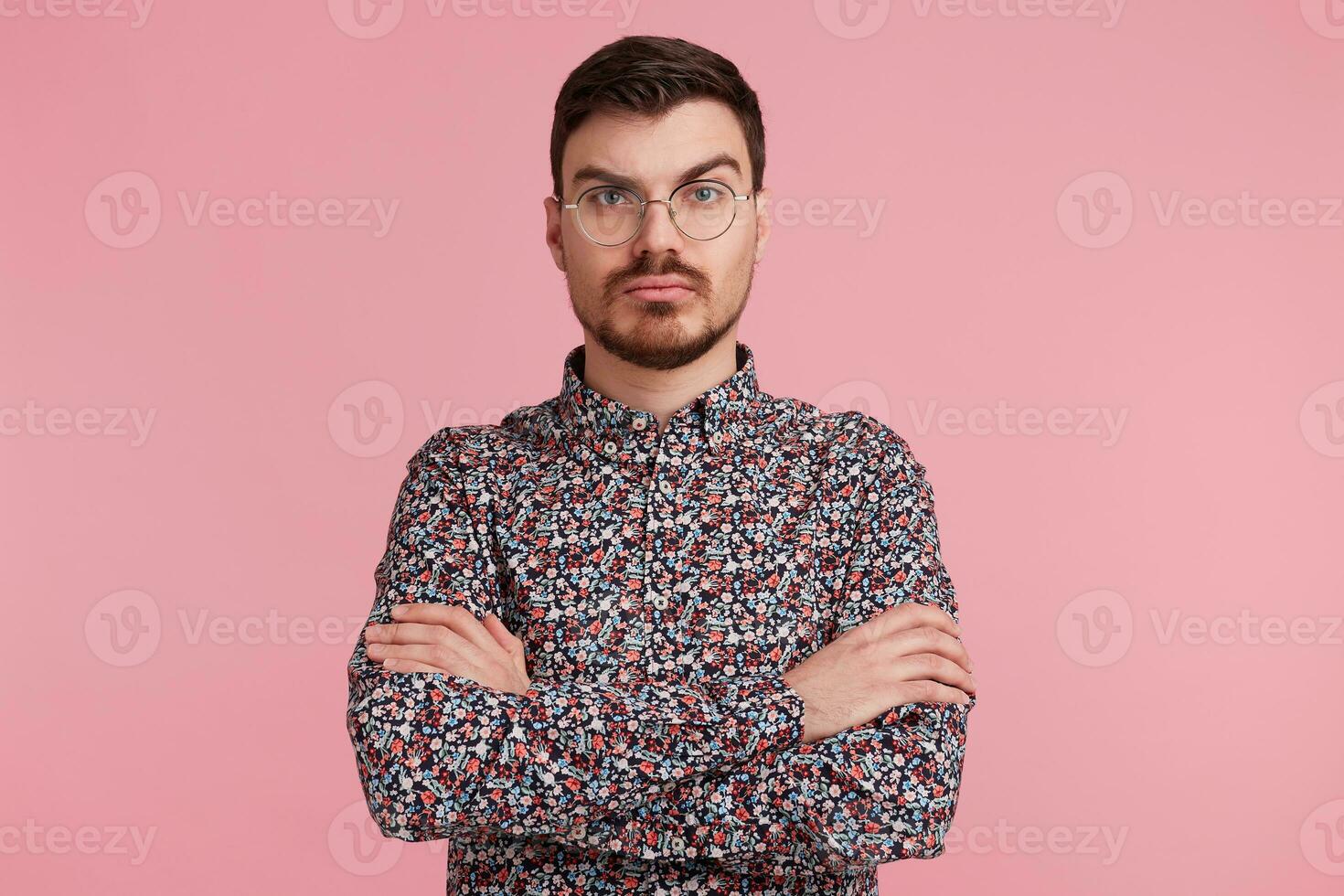 Pensive young bearded male wearing glasses in colorful shirt thinking over something, standing with hands crossed one brow raised questioning, having serious and puzzled expression over pink wall photo