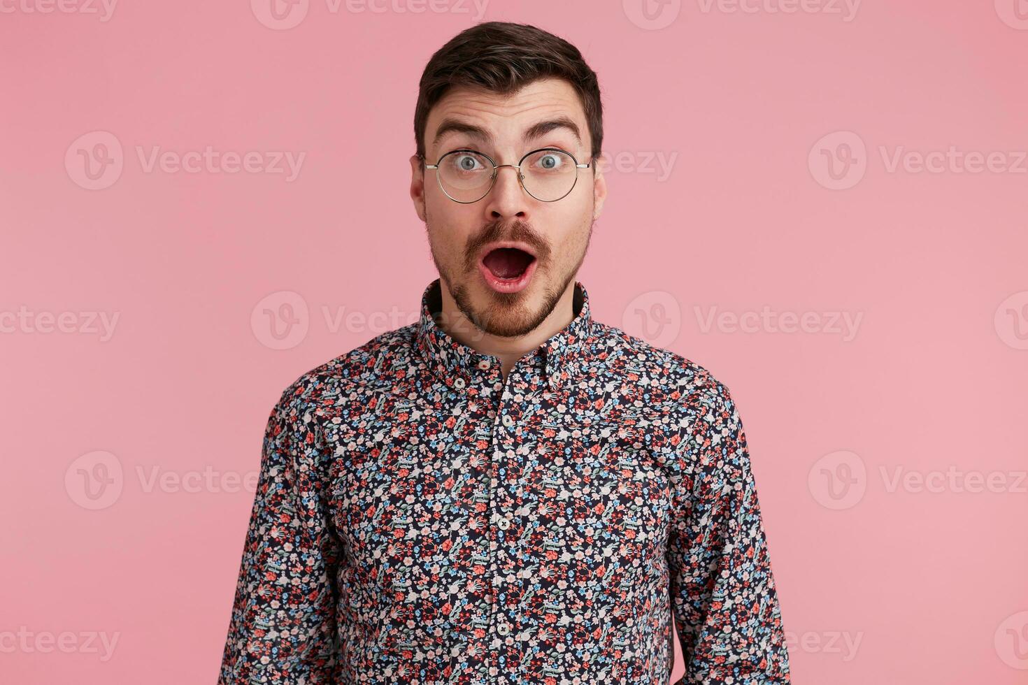Shocked attractive dark-haired handsome young man staring throught glasses, unshaved with beard and mustache in colorful shirt opened mouth from surprise, isolated over pink background photo