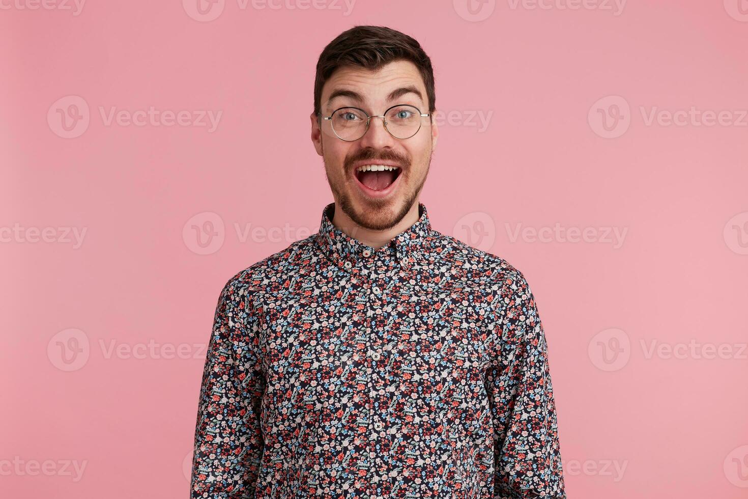 Portrait of a happy excited attractive handsome young man wearing glasses with dark hair unshaved with beard and mustache in colorful shirt opened mouth in amazement, isolated over pink background photo