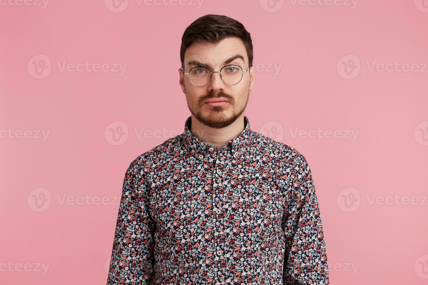 Portrait of suspicious pensive young bearded male wearing glasses in colorful shirt thinking over something, one brow raised questioning, having serious and puzzled expression over pink background photo