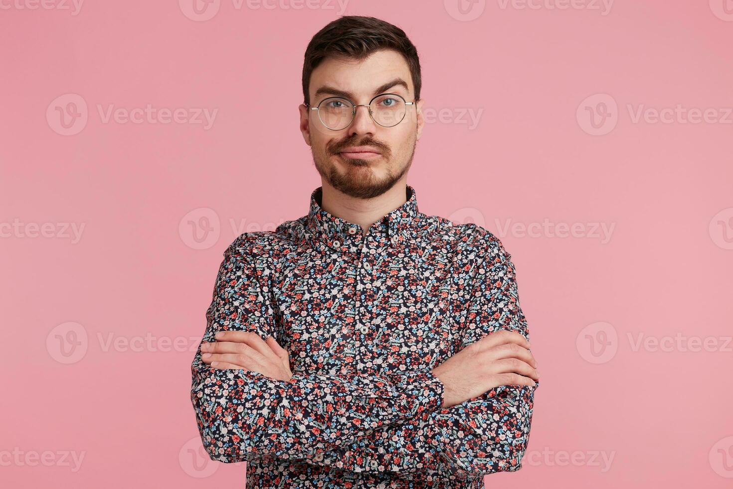 Looks skeptical young bearded male in glasses, colorful shirt thinking over something, standing with hands crossed one brow raised questioning, having perplexed and puzzled expression over pink wall photo