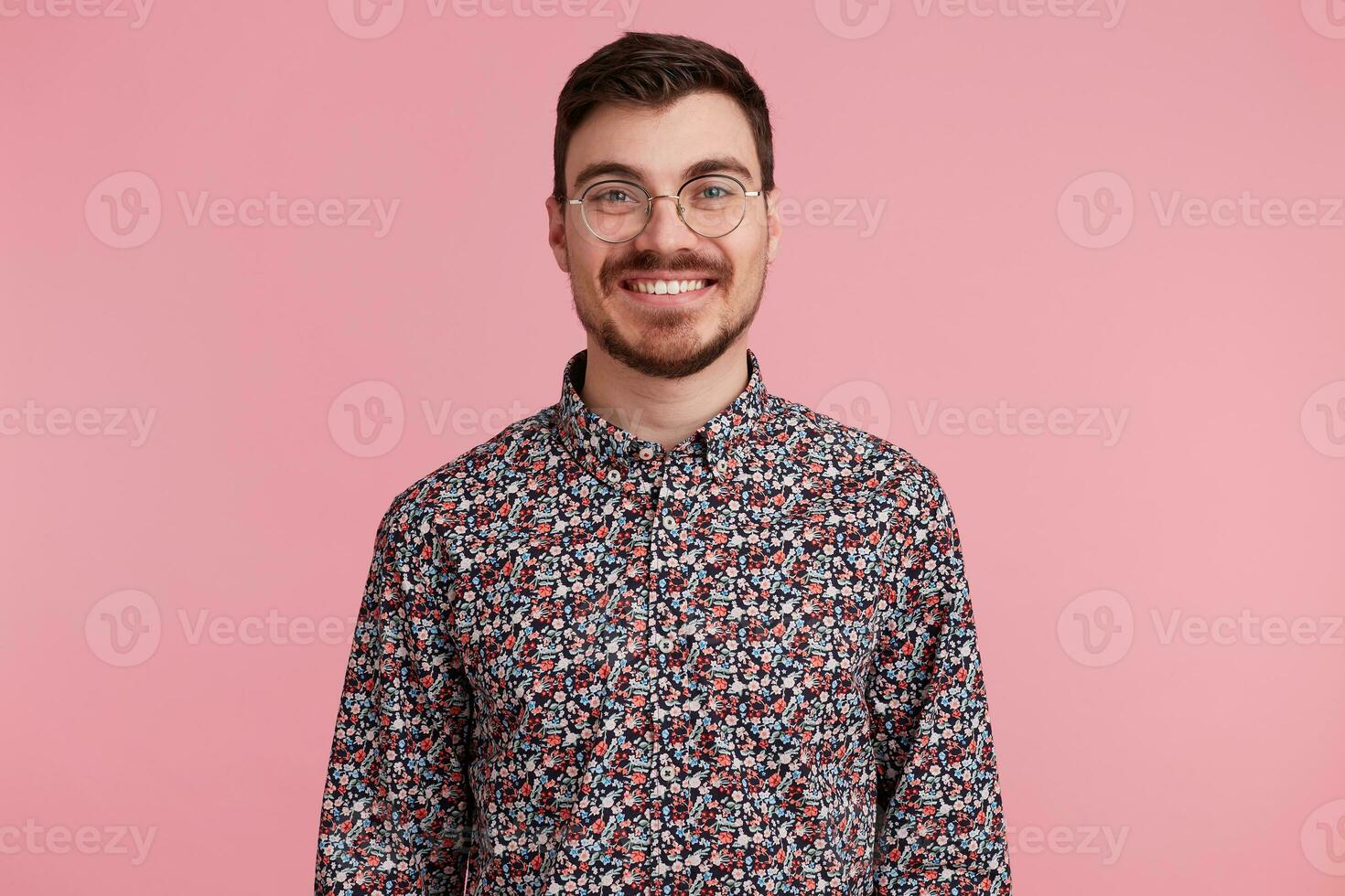 Portrait of a nice attractive handsome young man wearing glasses with dark hair unshaved with beard and mustache in colorful shirt pleasantly smiling, isolated over pink background photo