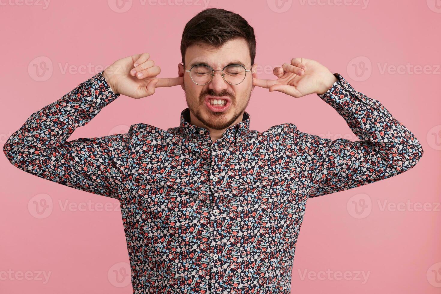 Close up of angry young bearded man dressed in colorful shirt, keeps closed eyes, two fingers close his ears, showing gesture of deafness, ignores someone, over pink wall photo