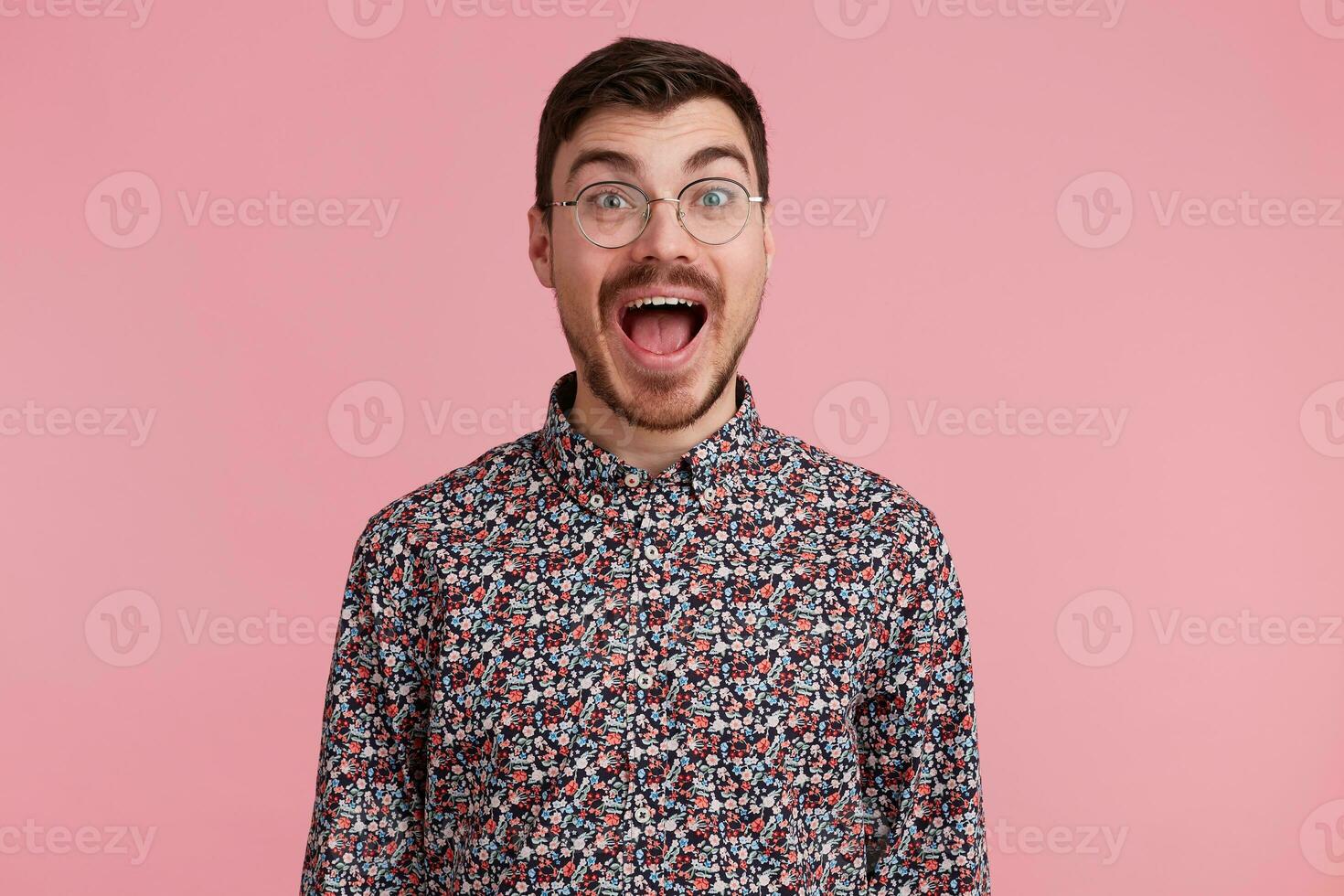 Excited man popping eyes and shouts loudly. Surprised male, keeps mouth open, being stunned, over pink background. Horizontal shot of shocked young male filled with emotion photo