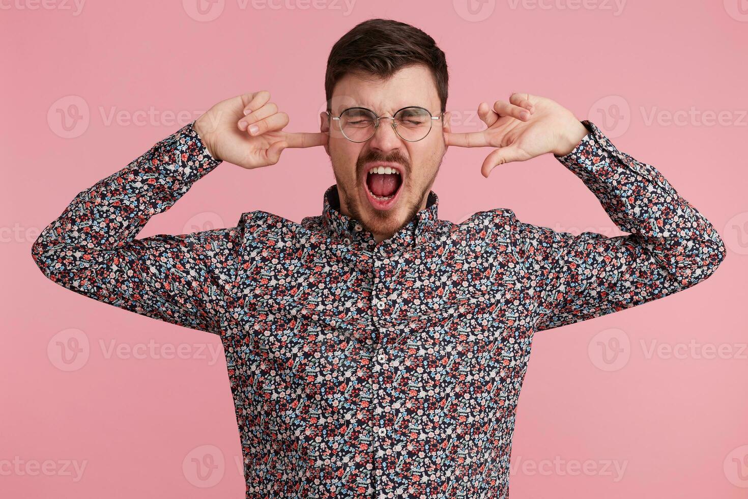 Close up of screaming shouting angry young bearded man dressed in colorful shirt, keeps closed eyes, two fingers close his ears, showing gesture of deafness, ignores someone, over pink wall photo