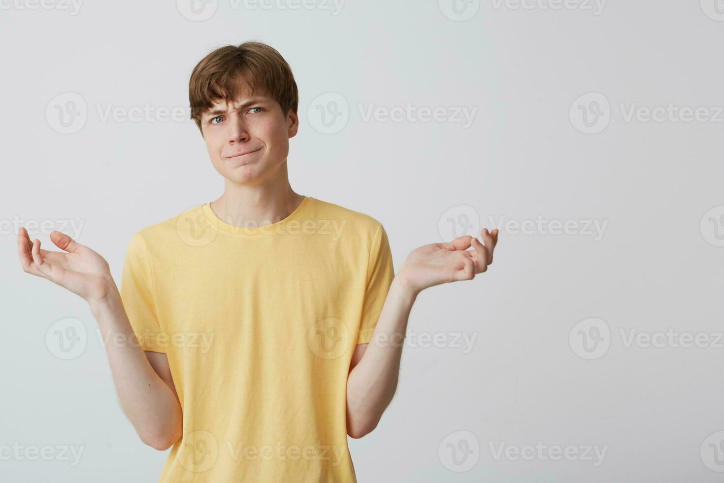 Portrait of confused upset young man wears yellow t shirt feels embarrassed and holding copyspace at both palms isolated over white background photo