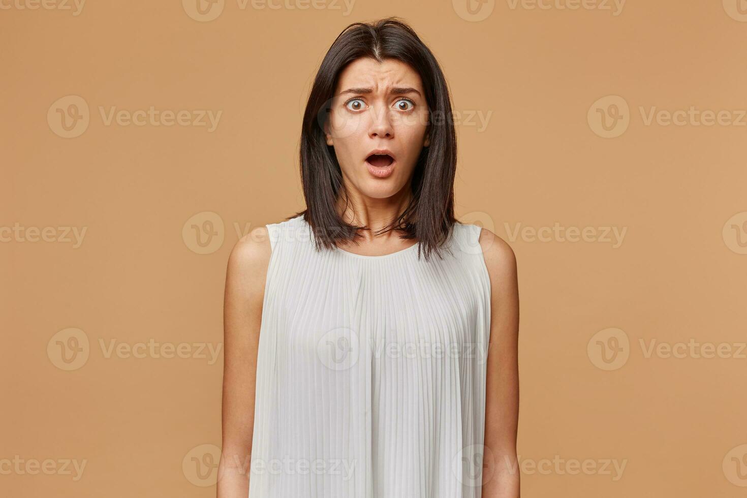 Portrait of scared woman in panic nervous frightened dressed in white dress, opened mouth feals fear, isolated on beige background photo