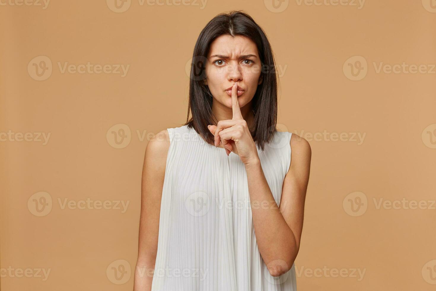 Anxious, worried, worried, nervous, panicked, aggressive, angry woman demonstrates a gesture of silence, holding an index finger near the mouth calls for privacy to be kept, secret, over beige wall photo