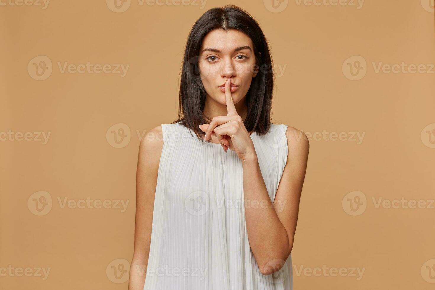 Mysterious girl in dress with calm facial expression, demonstrates a gesture of silence, holding an index finger near the mouth calls for privacy to be kept, secret, isolated over beige background photo