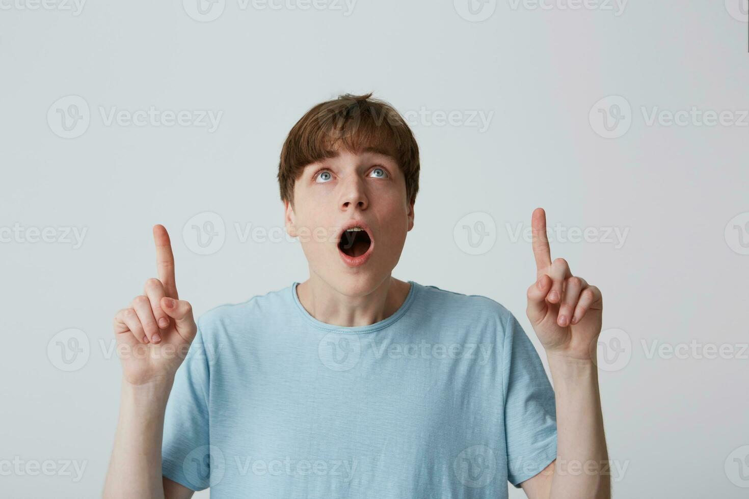 Portrait of surprised astonished young man with opened mouth wears blue t shirt feels surprised and points up to copyspace with both hands isolated over white background photo