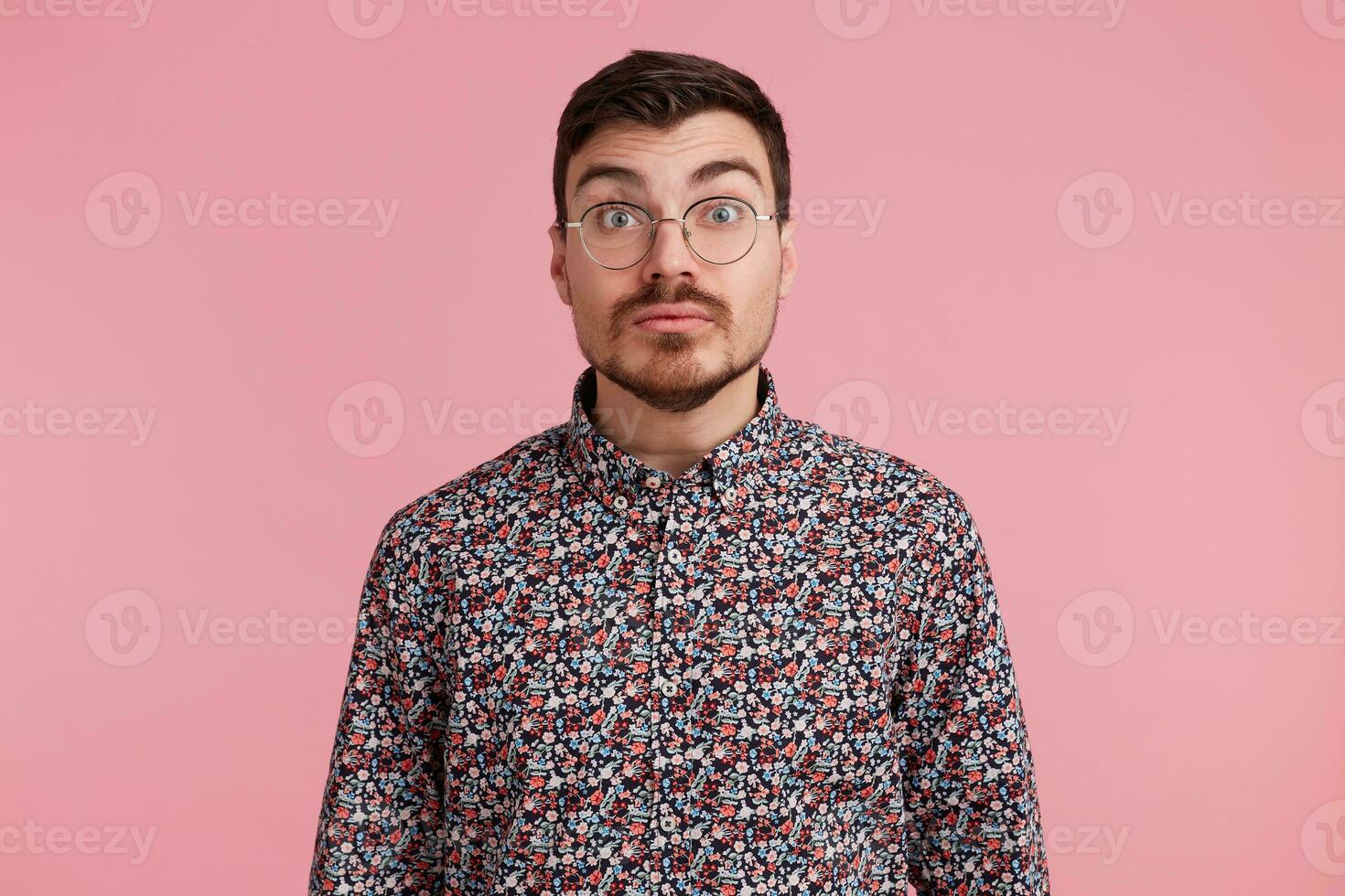 Shocked uncertain surprised man stares through spectacles wearing colorful shirt tries to make situation clear, over pink background photo