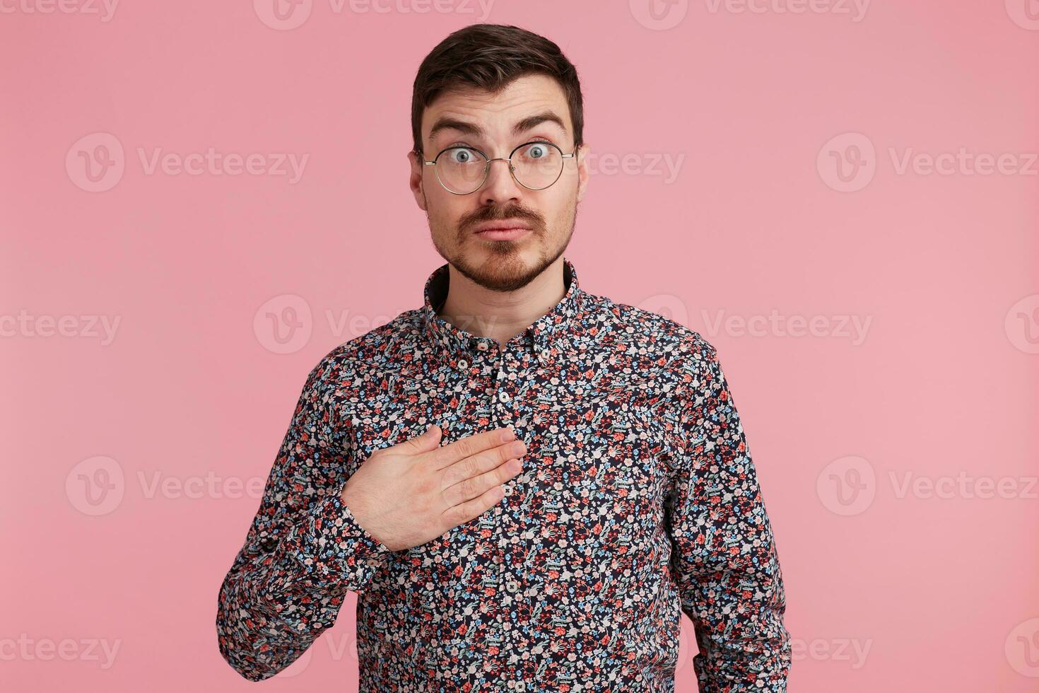 Surprised uncertain surprised man stares through spectacles wearing colorful shirt pointin with palm on himself tries to make situation clear, over pink background photo