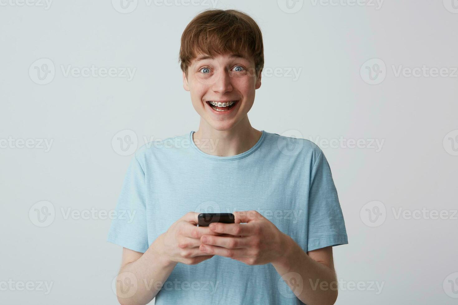 Closeup of cheerful excited young man with braces on teeth wears blue t shirt feels happy and using smartphone isolated over white background photo