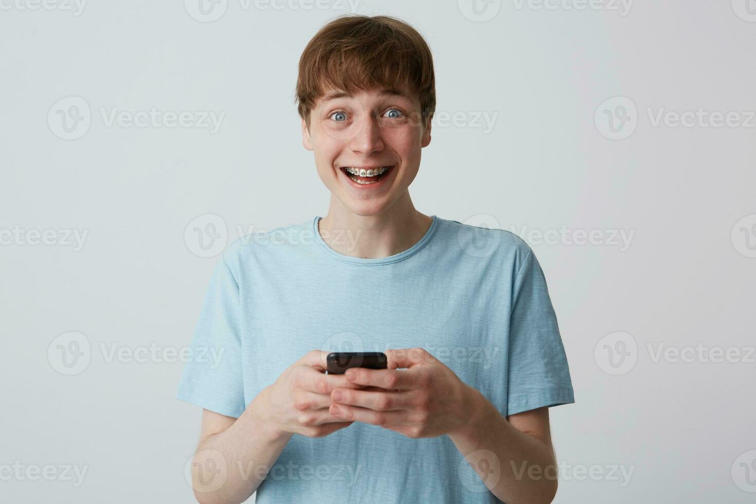 de cerca de alegre emocionado joven hombre con tirantes en dientes usa azul t camisa siente contento y utilizando teléfono inteligente aislado terminado blanco antecedentes foto