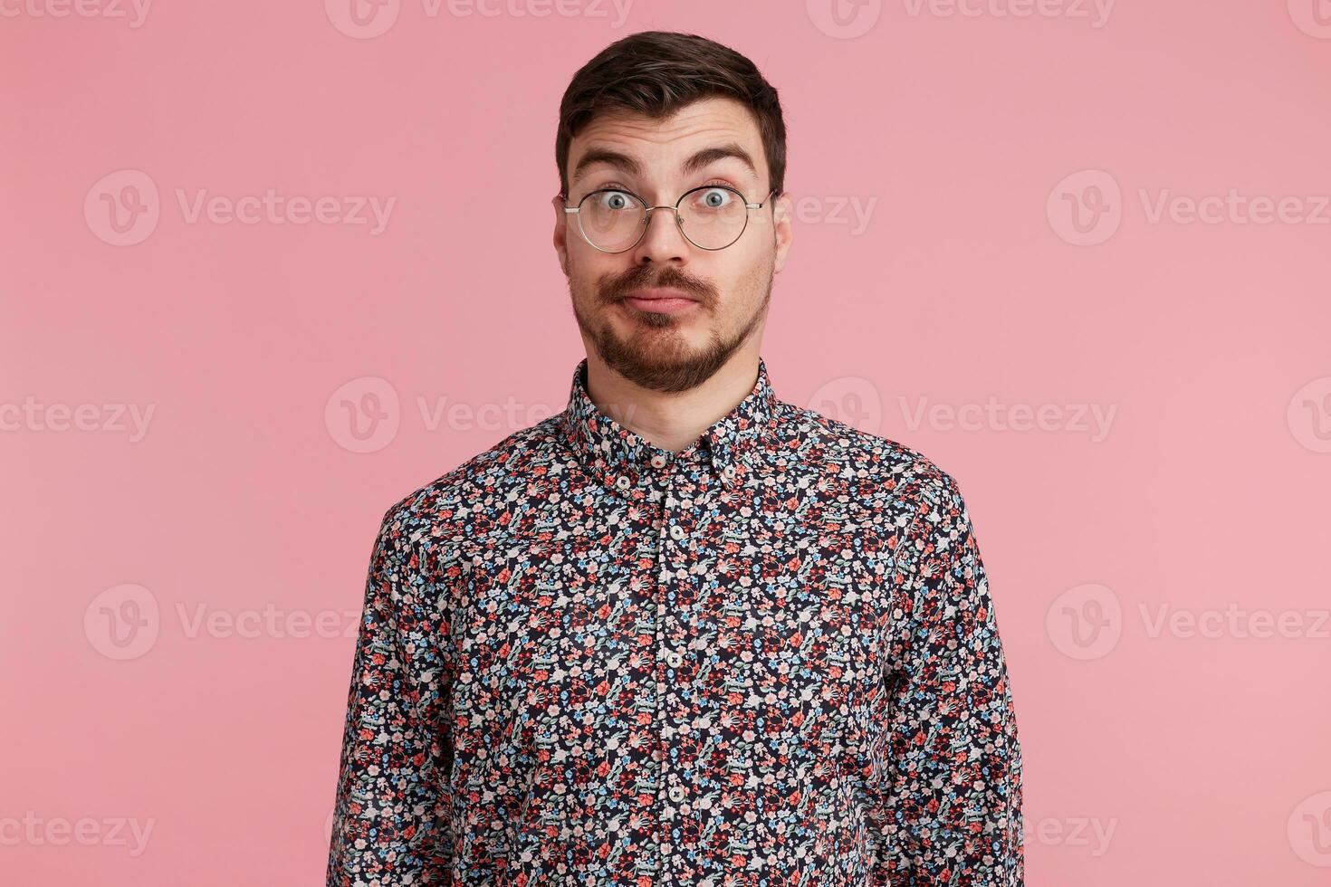 Surprised man stares through spectacles with misunderstanding, bewilderment, wearing colorful shirt shrug shoulders in uncertainty, over pink background photo