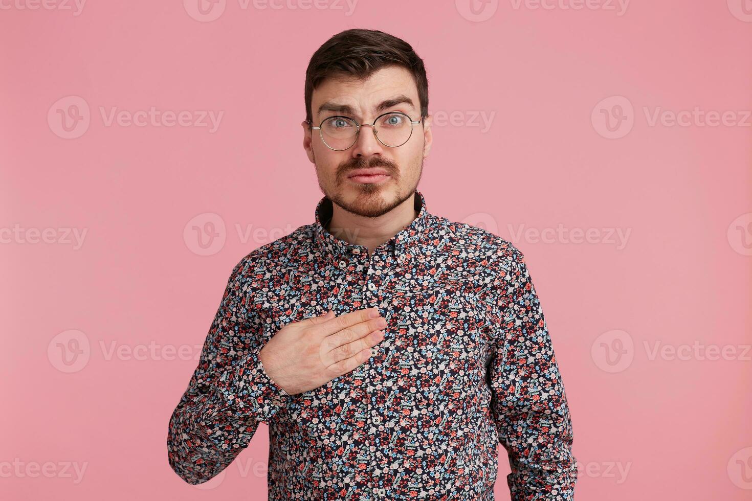 Uncertain surprised man in spectacles wearing colorful shirt pointin with palm on himself tries to make situation clear, over pink background photo