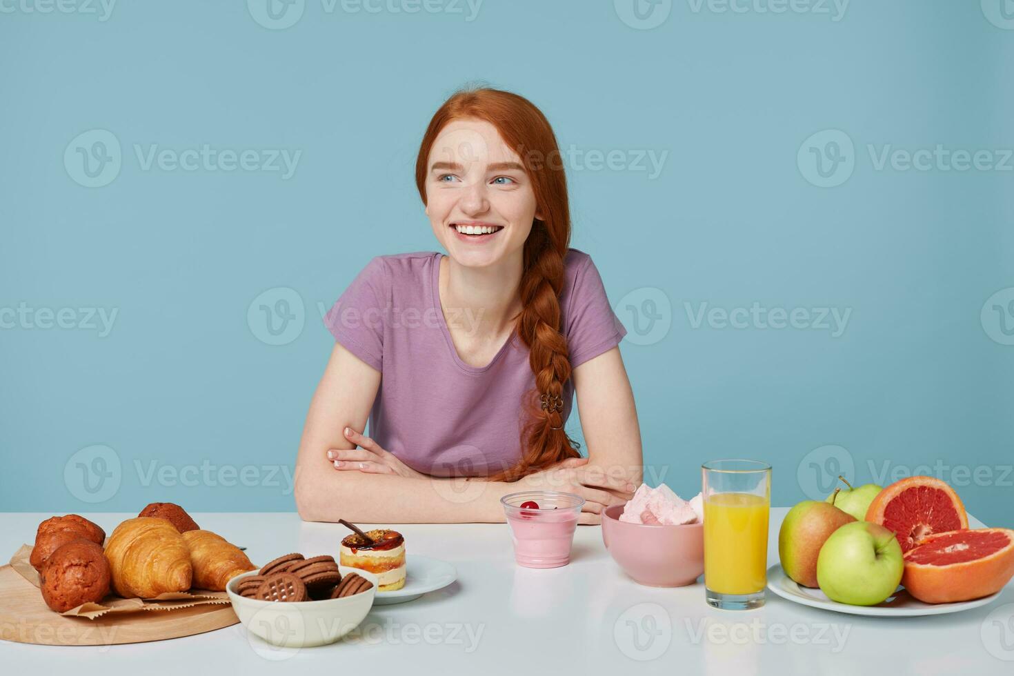 Studio shot of smiling red-haired girl with braided hair sitting at a table, about to eat lunch looking leftside. On the white table lay baking and fresh healthy food. photo