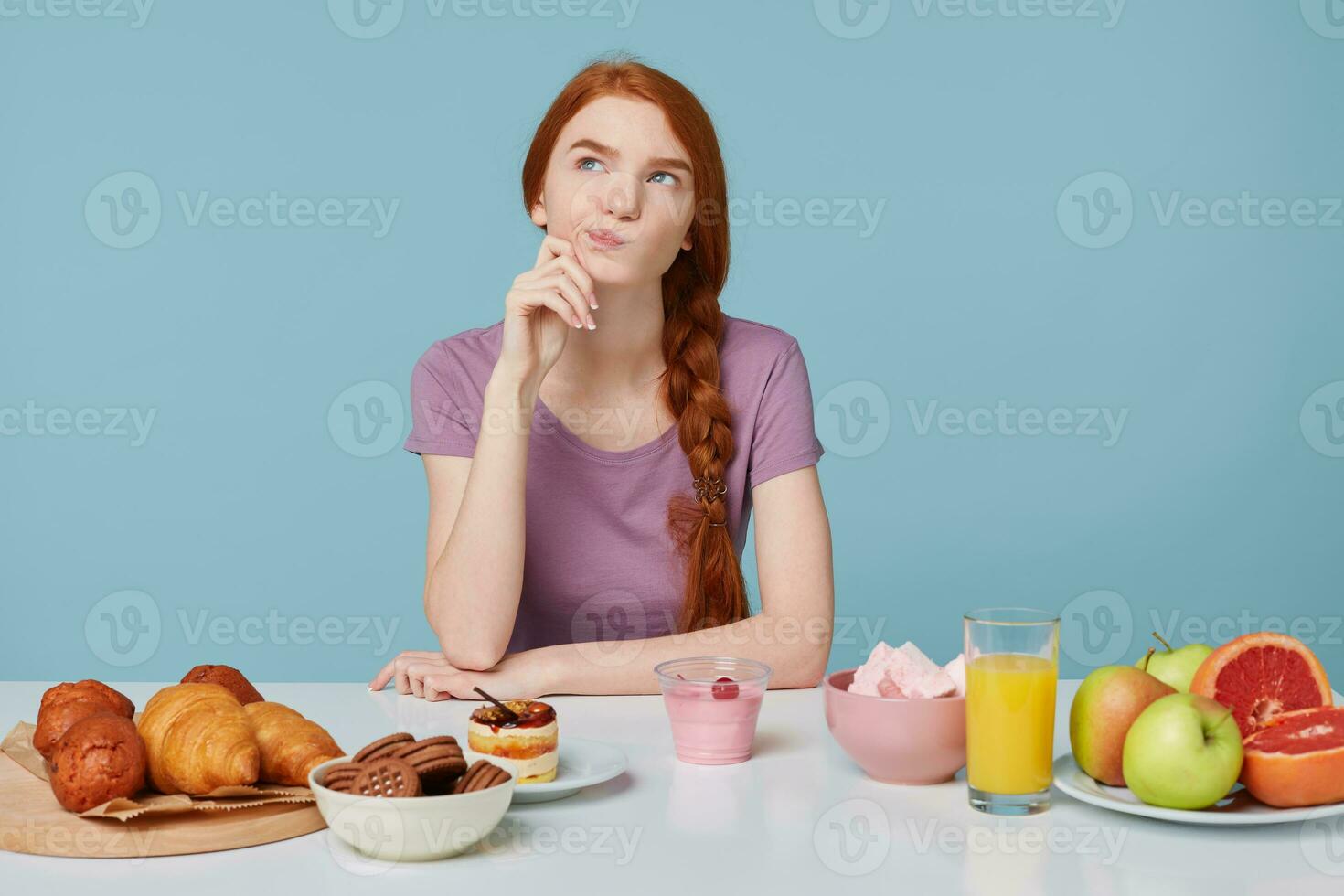 Photo of red-haired girl looking upwards with dissatisfaction, doubts thinks about diet,health, nutrition, extra calories,baking food and fresh fruit juice yogurt lay on the table,on a blue background