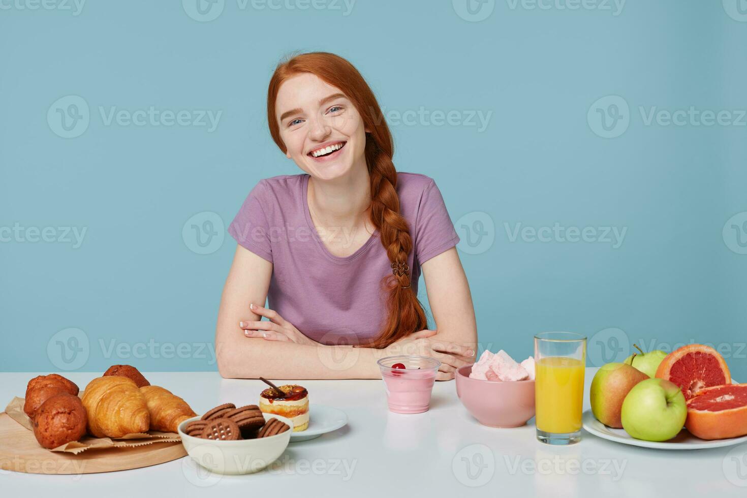 Young attractive red-haired girl smiles dreamily looks to the upper right corner, sitting at a white table on which are fresh, healthy products and delicious pastries baking products. photo