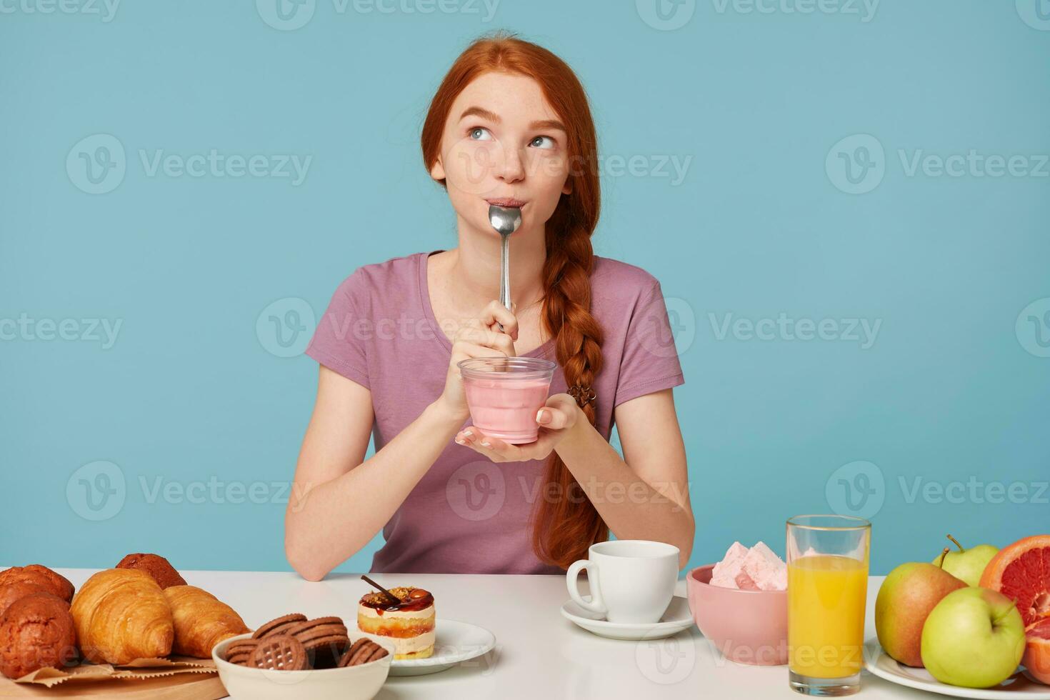 Nice red-haired girl with a braid sits at a table, has lunch, holds in her hands a glass, dreamingly looks at left upper corner and licks teaspoon with pleasure after tasting yummy yogurt. photo