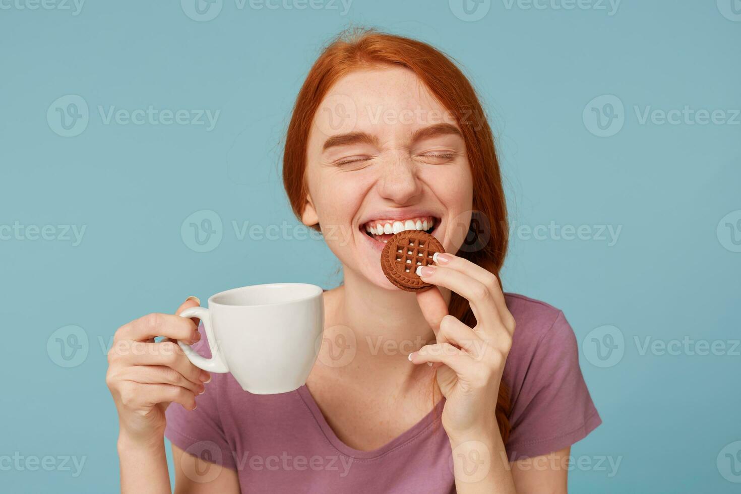 Close up of a lovely beautiful red-haired girl with closed eyes,feels pleasure, keeps in hand white cup with drink, with appetite bites yummy chocolate cookie, over blue background photo