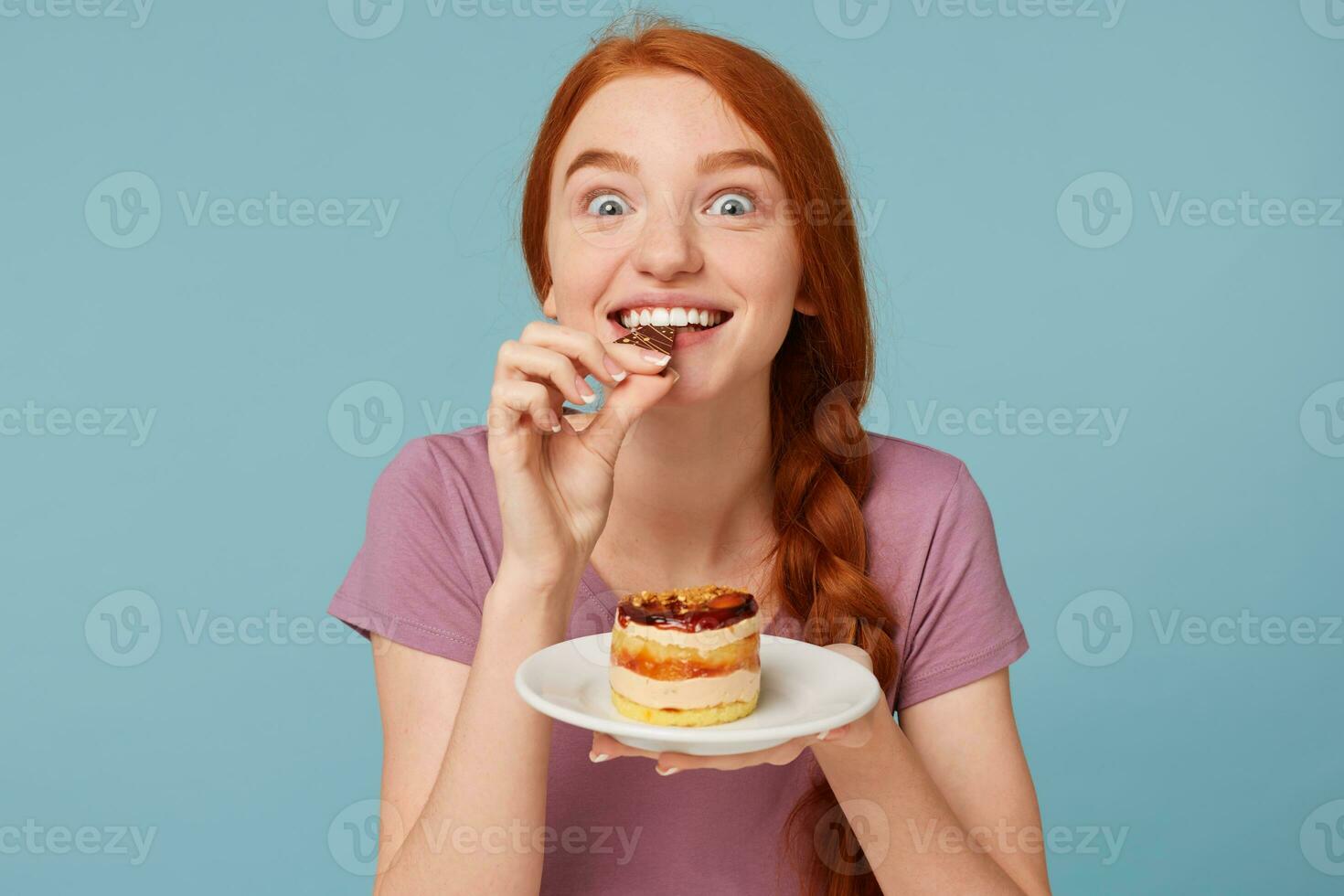 Close up of a red-haired girl eagerly tastes a delicious cake, bites off a piece of chocolate and holds a plate in her hands isolated on a blue background. photo