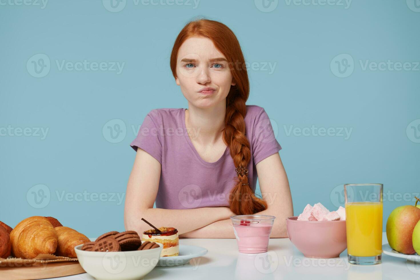 Photo of red-haired girl looking camera with anger discontent, doubts thinks about diet, extra calories, baking food and fresh fruit juice yogurt lay on the  table, isolated on a blue background