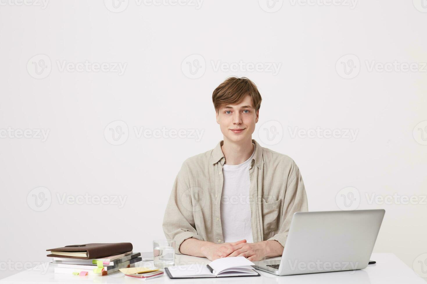 Portrait of handsome young man student wears beige shirt sitting at the table with laptop computer and notebooks isolated over white background photo