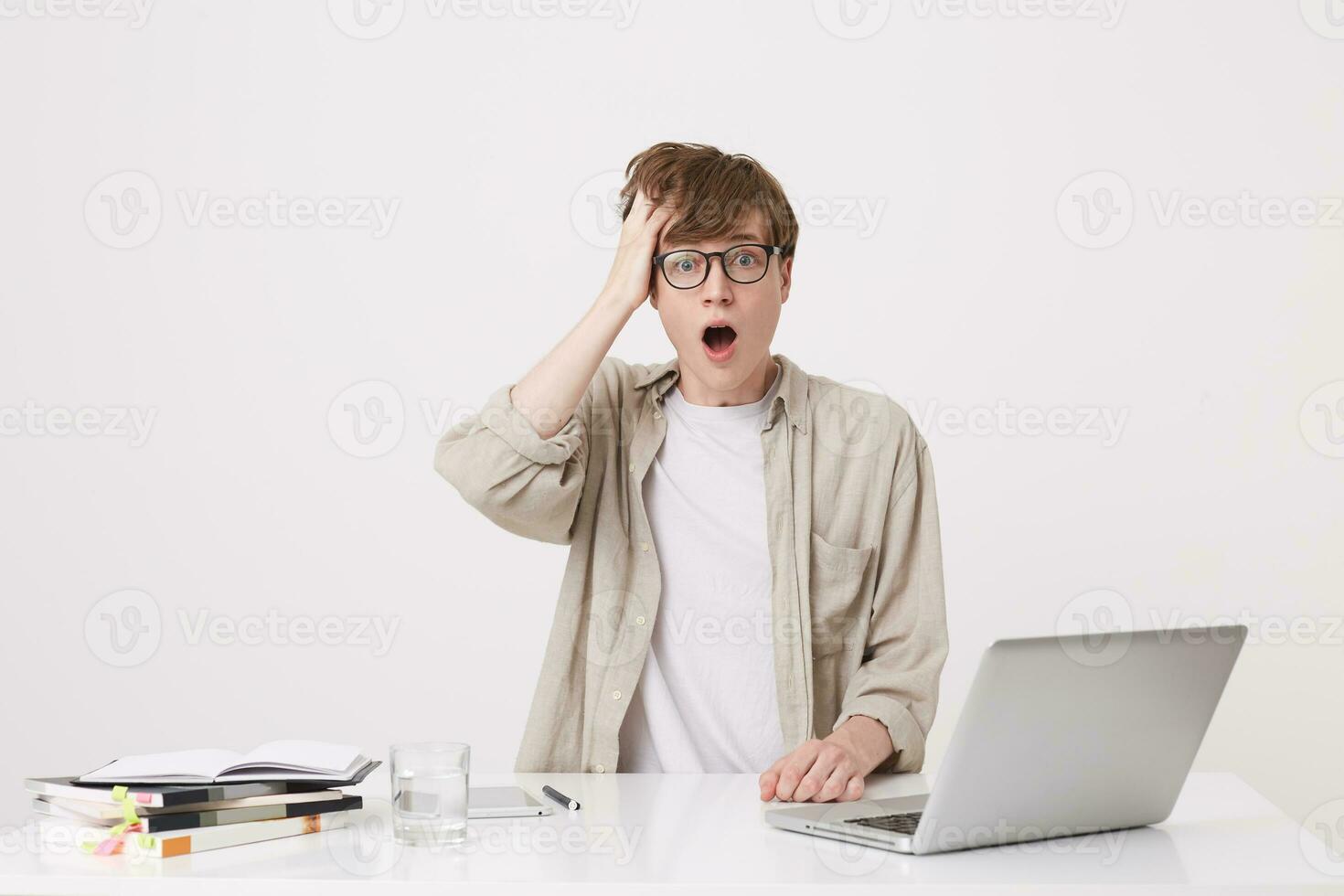 Closeup of amazed young man student with opened mouth wears beige shirt and glasses looks shocked sitting with laptop computer and notebooks at the table isolated over white background photo