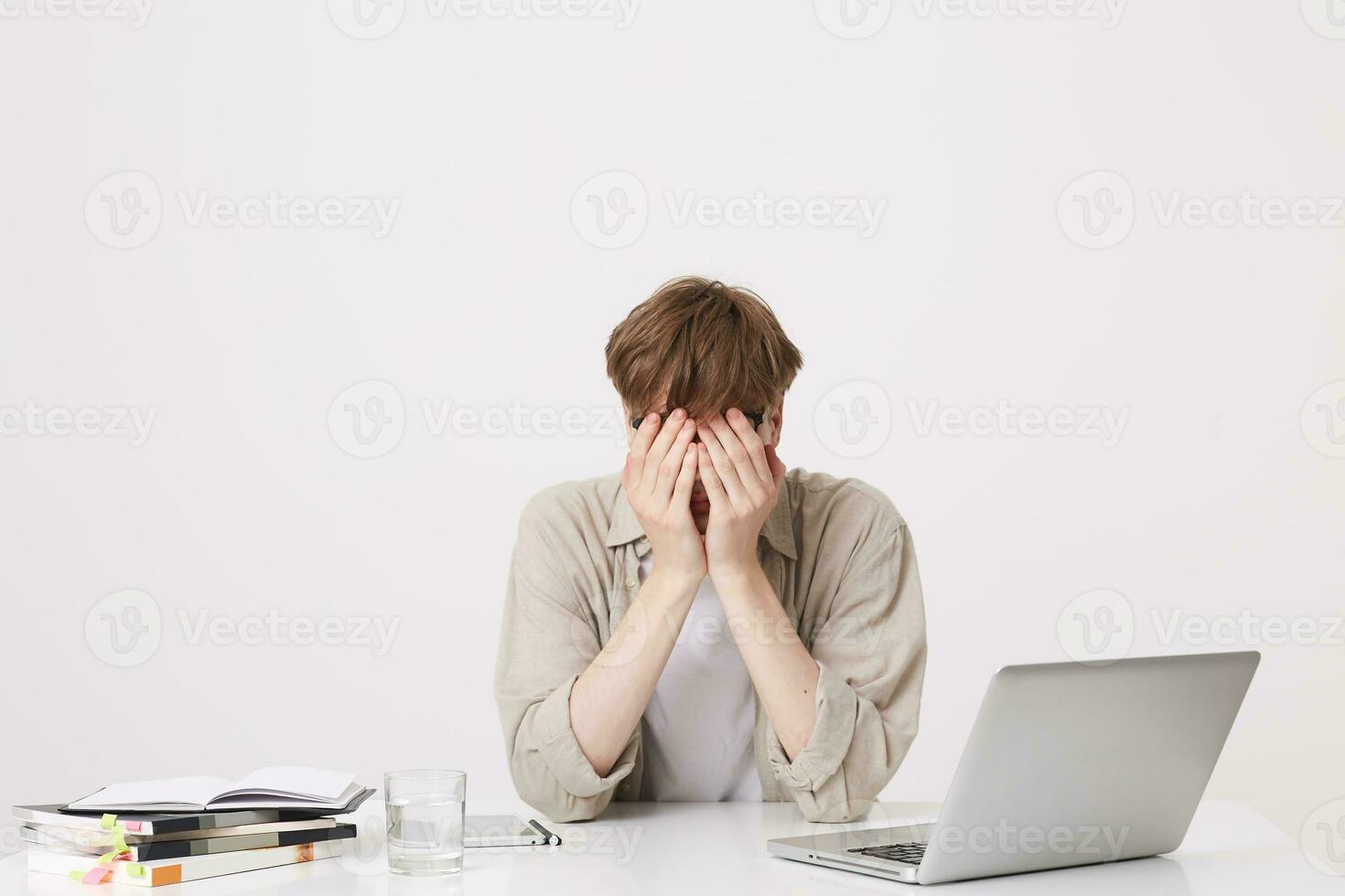 Closeup of tired desperate young man student wears beige shirt coveres face by hands sitting at the table with laptop computer and notebooks isolated over white background photo