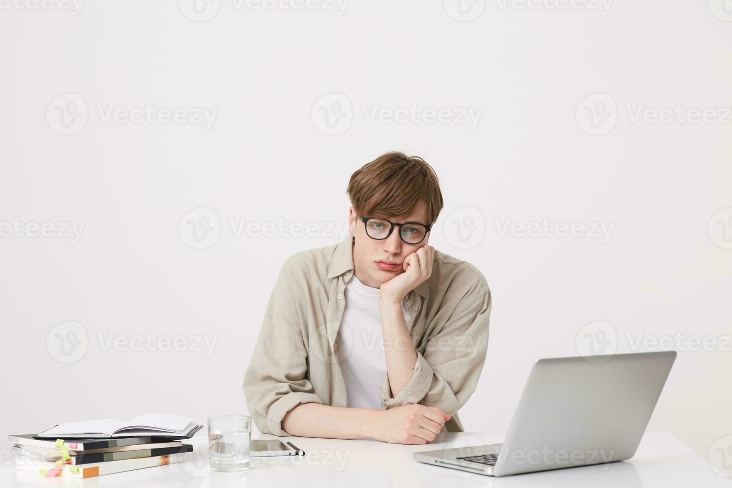 Portrait of upset depressed young man student wears beige shirt and spectacles looks bored and study at the table with laptop computer and notebooks isolated over white background photo