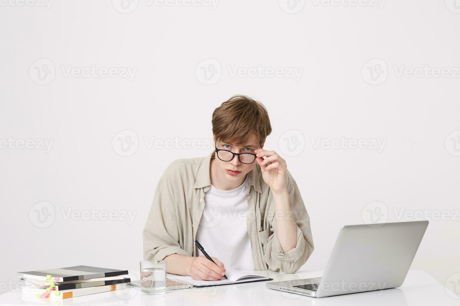 Portrait of concentrated young man student wears spectacles and beige shirt writing and study at the table with laptop computer and notebooks isolated over white background photo