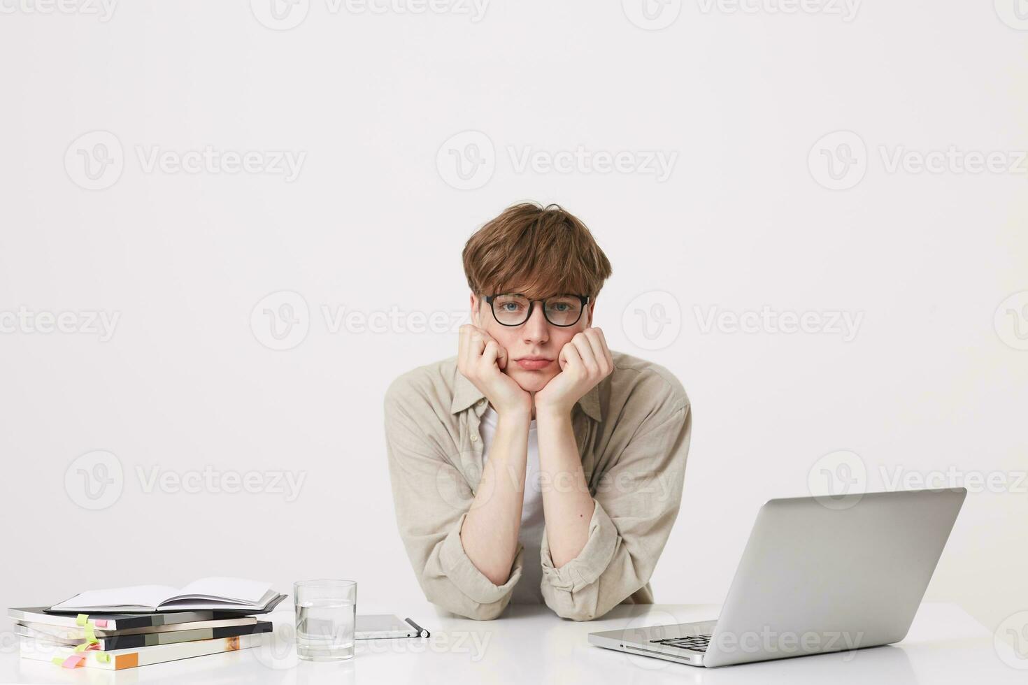 Portrait of upset unhappy young man student wears beige shirt and glasses feels sad and study at the table with laptop computer and notebooks isolated over white background photo