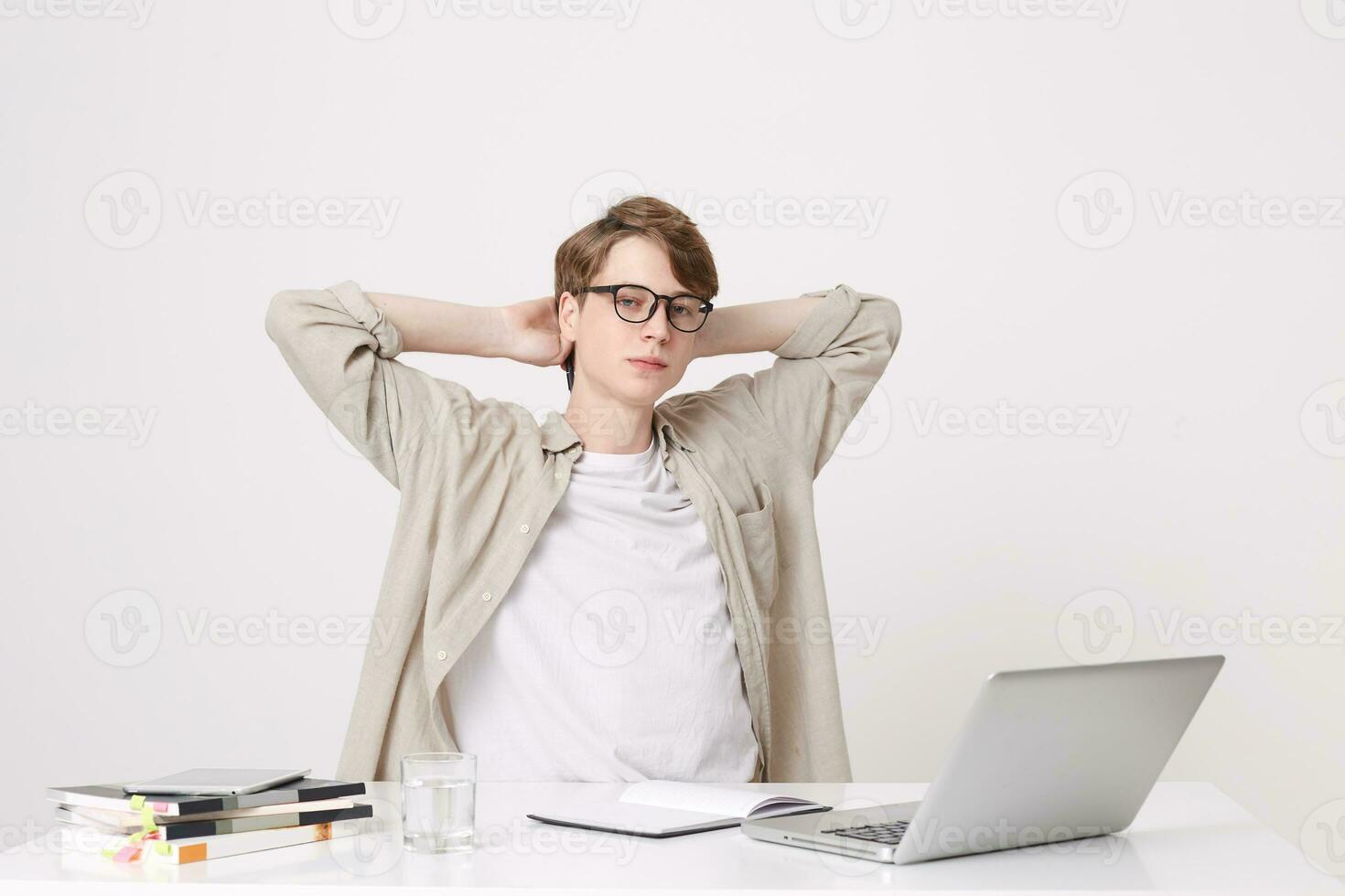 Closeup of relaxed confident young man student wears beige shirt and glasses sitting with hands over head at the table with laptop computer and notebooks isolated over white background photo