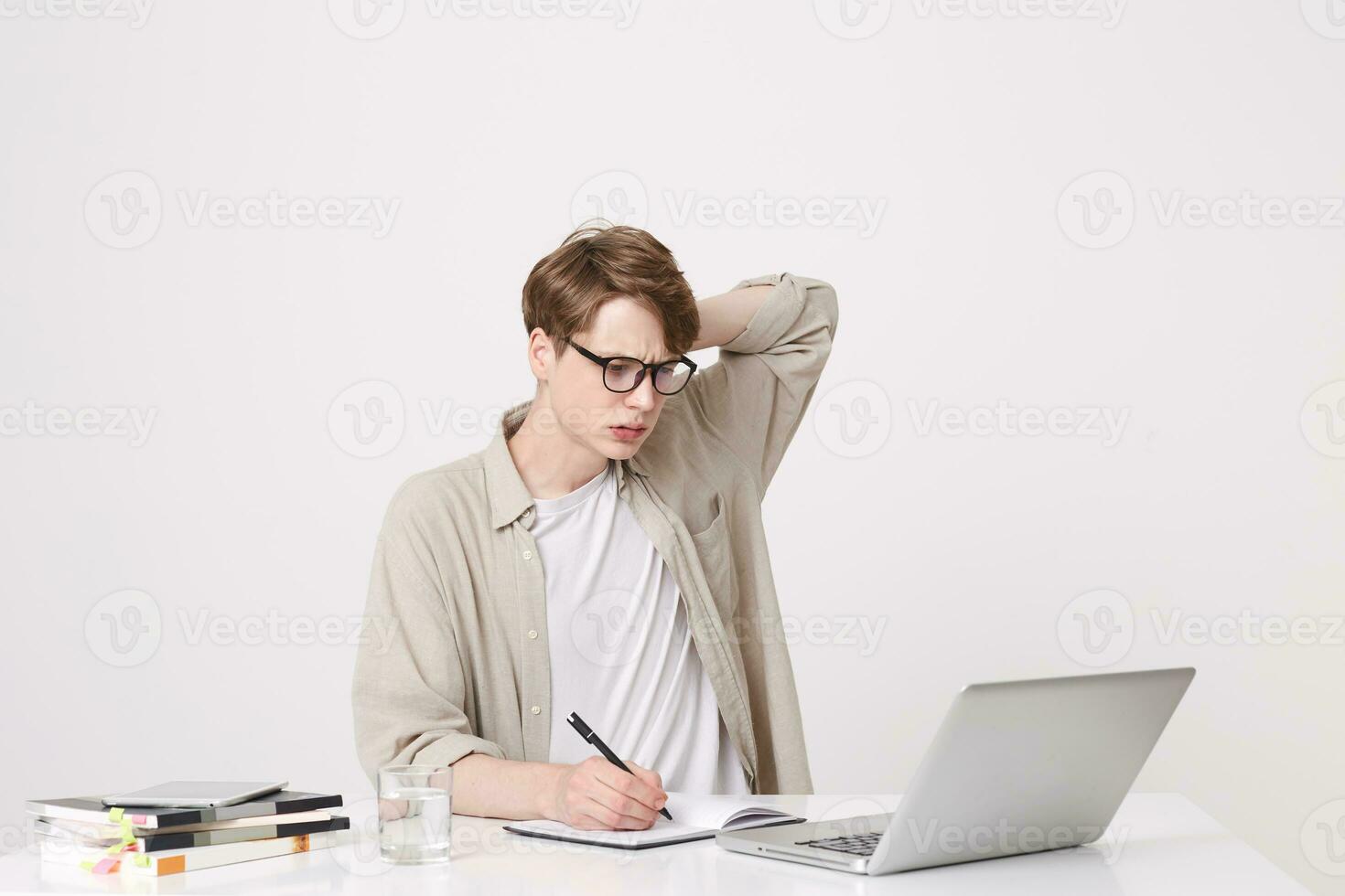 Portrait of thoughtful young man student wears beige shirt and spectacles looks concentrated and study at the table using laptop computer and notebooks isolated over white background photo