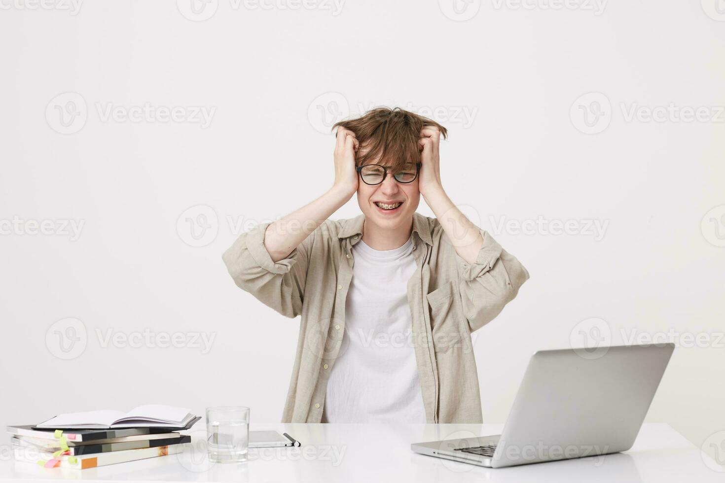 Portrait of mad crazy young man student wears beige shirt and spectacles feels irritated and study at the table with laptop computer and notebooks isolated over white background photo