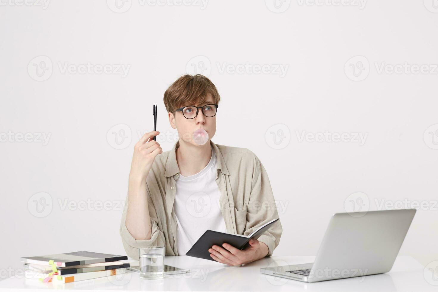 retrato de pensativo joven hombre estudiante usa beige camisa y lentes pensando y soplo burbujas con masticación goma a el mesa con ordenador portátil computadora y cuadernos aislado terminado blanco antecedentes foto