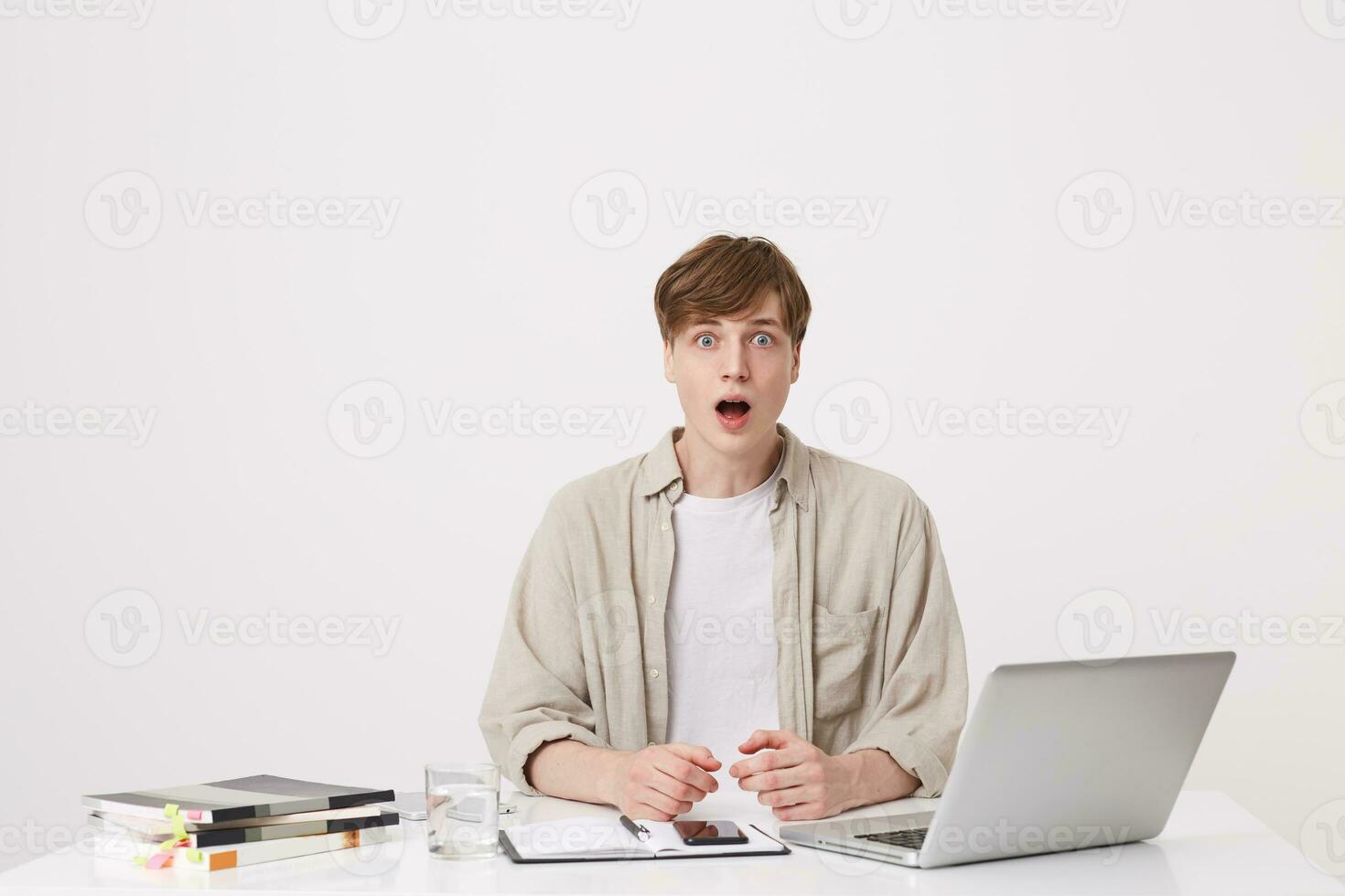 Closeup of stunned young man student with opened mouth wears beige shirt sitting at the table and using laptop computer and notebooks isolated over white background photo