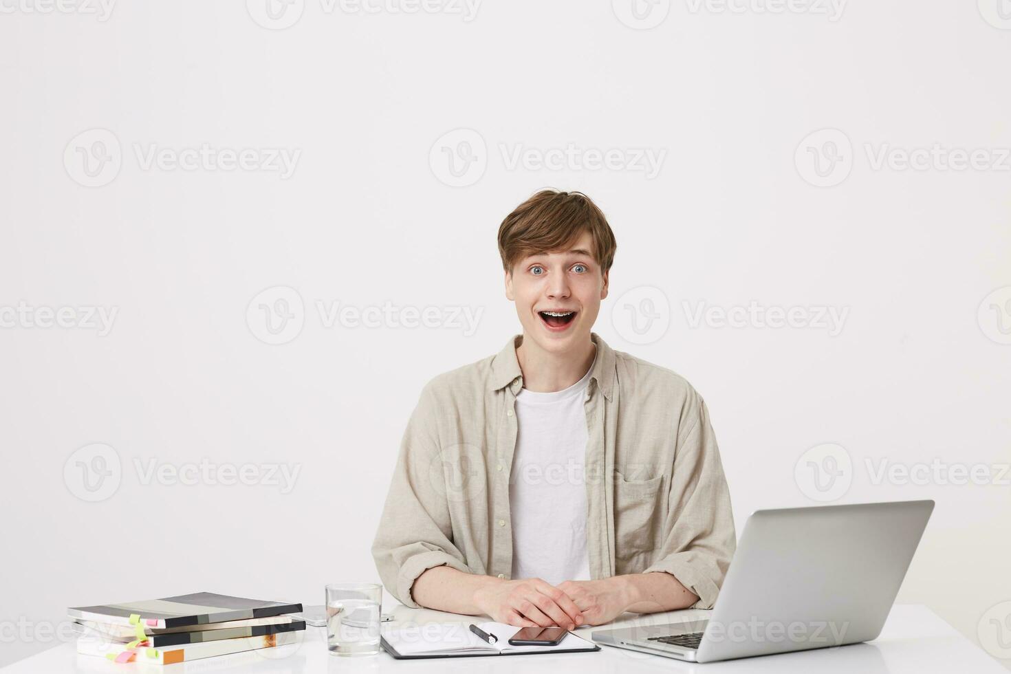 Closeup of surprised young man student with braces wears beige shirt feels impressed and using laptop computer and notebooks at the table isolated over white background photo