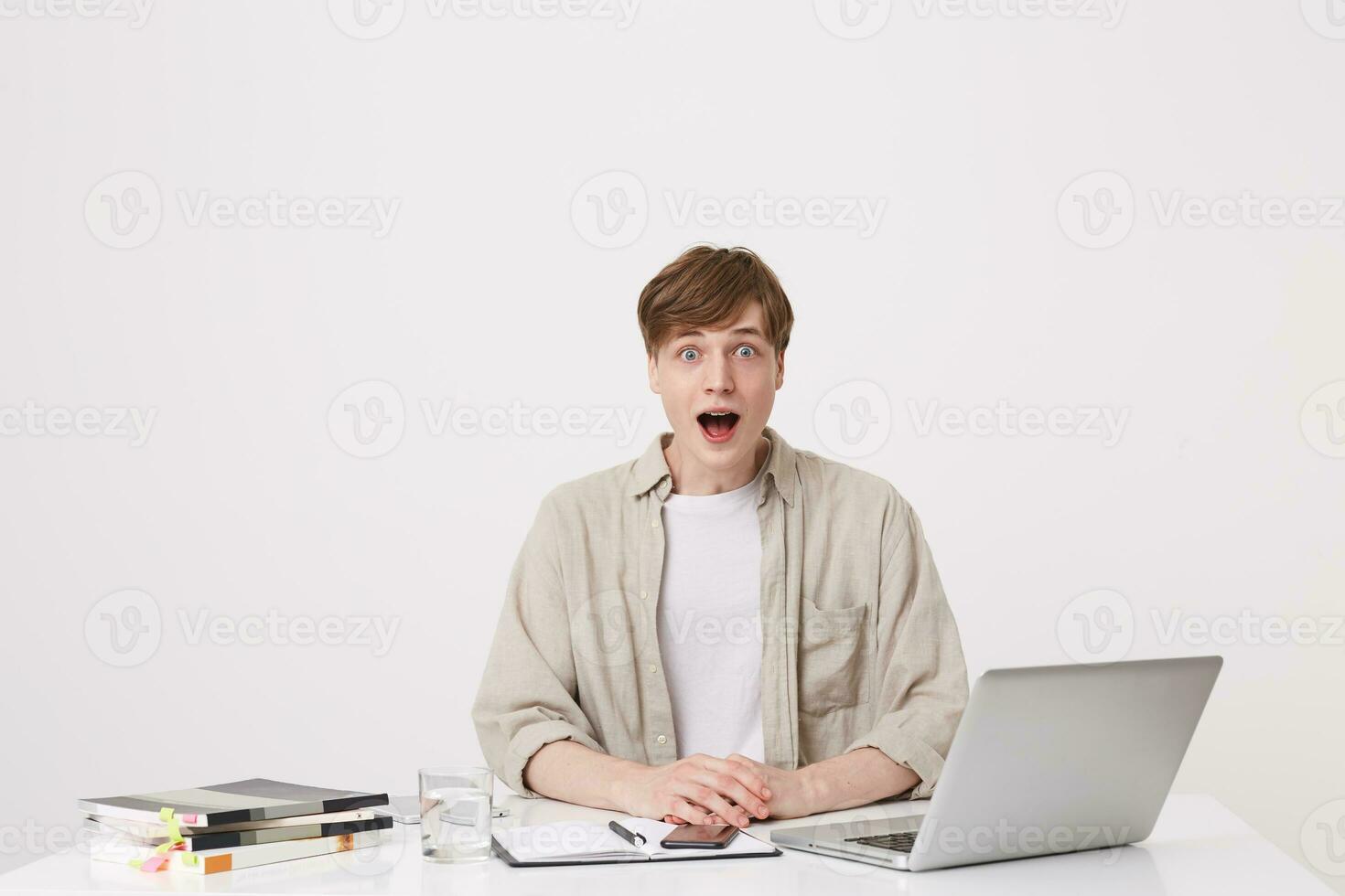 Portrait of shocked young man student opened mouth wears beige shirt feels astonished and using with laptop computer and notebooks at the table isolated over white background photo