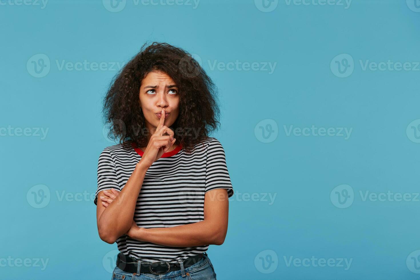 Young afro american woman with afro hairstyle wearing stripped t-shirt over isolated blue background with copy space thinking pondering about something concentrated with finger on lips. photo