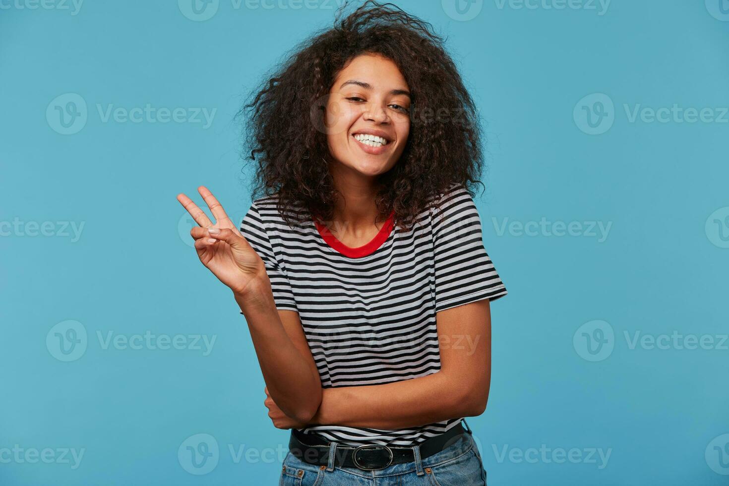 African American pretty smiling girl with afro hairstyle looking at camera and doing peace sign with hand or V sign with fingers. Isolated over blue background, copy space photo