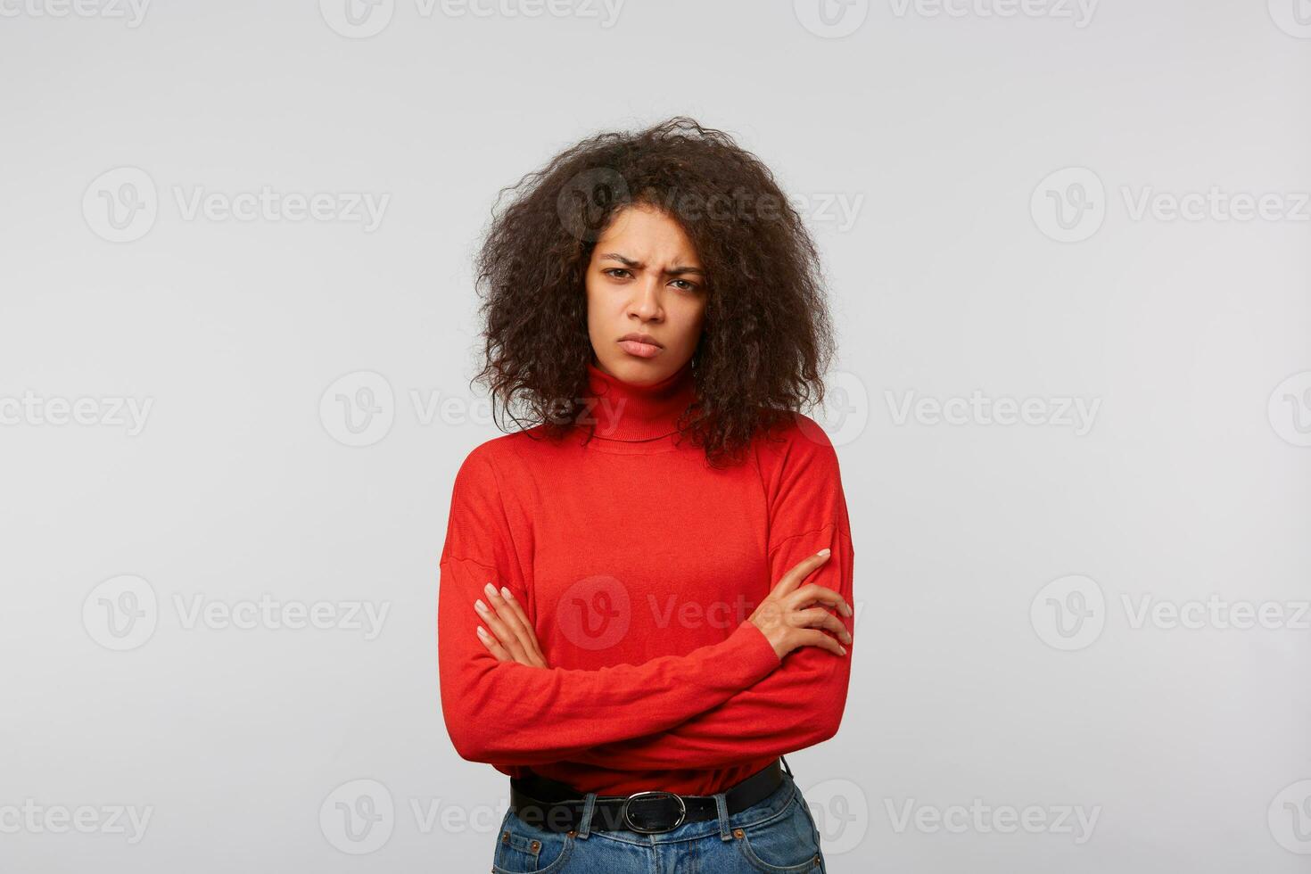Serious suspicious latino woman with curly afro hair standing with crossed hands and frowning, expressing distrustfulness and disappointment, over white background photo