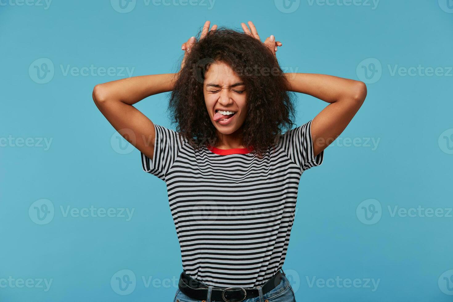 Close up of funny joking beautiful young woman, feels happy, makes horns on her head with fingers, shows tongue, closed eyes, isolated over blue background photo