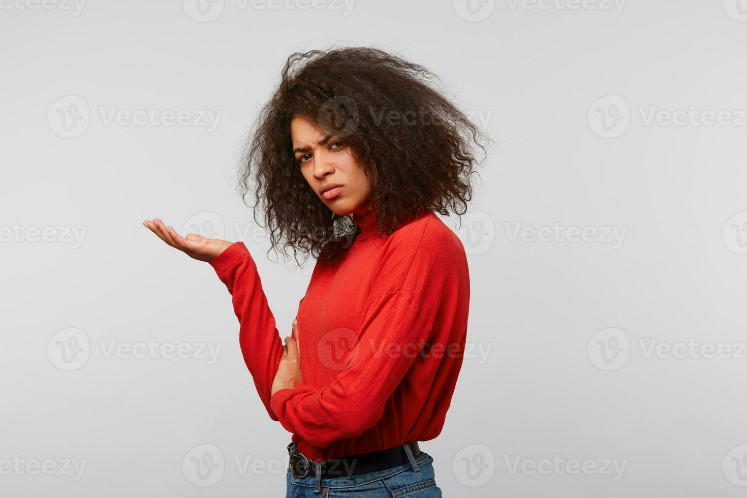 ofendido insatisfecho irritado latino mujer con afro peinado en un rojo manga larga en pie oblicuo con palma arriba en un blanco antecedentes foto