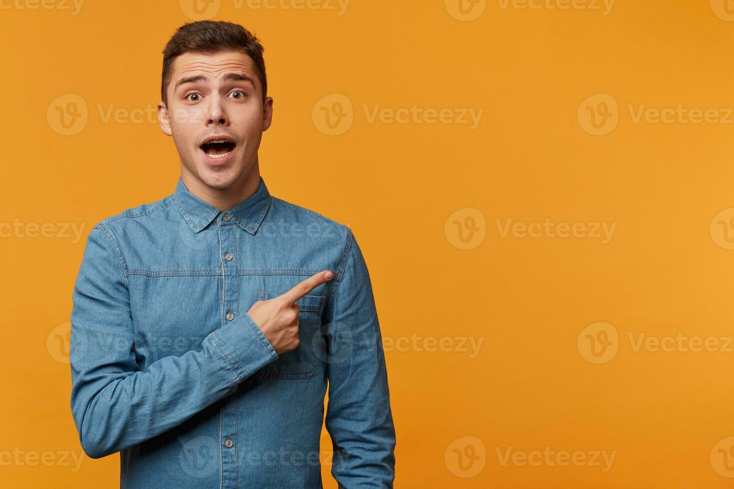 Man in panic nervous excited dressed in denim shirt pointing right with his finger, feals fear, isolated on yellow background with copy space for your text or advertisement photo