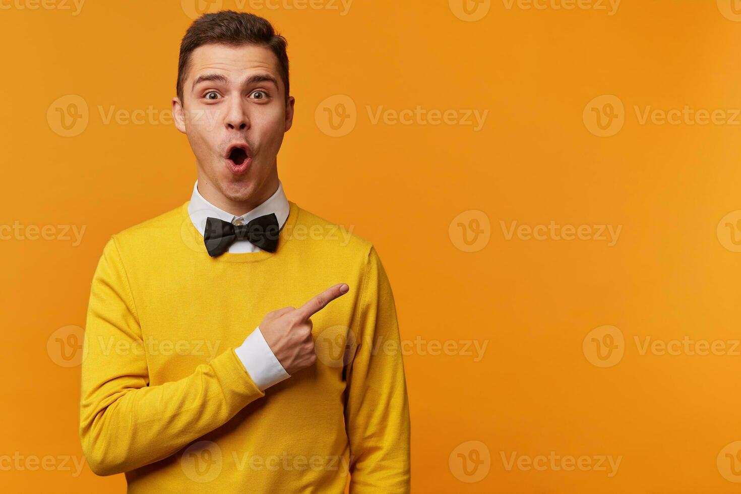 Young surprised man in yellow sweater over white shirt and black bow-tie pointing right with his finger, shouting WOW, isolated on yellow background with copy space for your text. Shock content photo